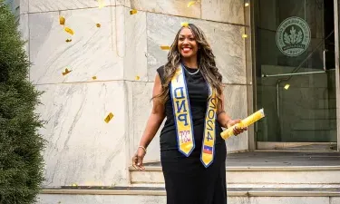 Graduating student celebrating with confetti, wearing a black dress and DNP 2024 sash, holding a diploma outside a building.