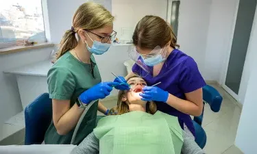 Two dental professionals in protective gear and masks performing a dental procedure on a patient in a modern dental office, showcasing teamwork and hygiene practices.